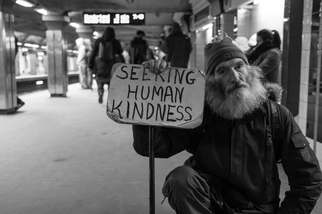 Man who is homeless holding sign that says "Seeking Human Kindness"